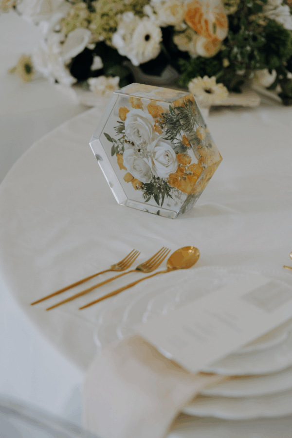 Elegante Tischdekoration mit hexagonalem Acrylblock, in dem weiße Rosen und apricotfarbene Blüten in Kunstharz konserviert sind. Im Hintergrund ein passender Hochzeitsstrauß, im Vordergrund goldenes Besteck auf feinem weißen Tischtuch - perfekte Hochzeitserinnerung als stilvolles Dekorationselement.