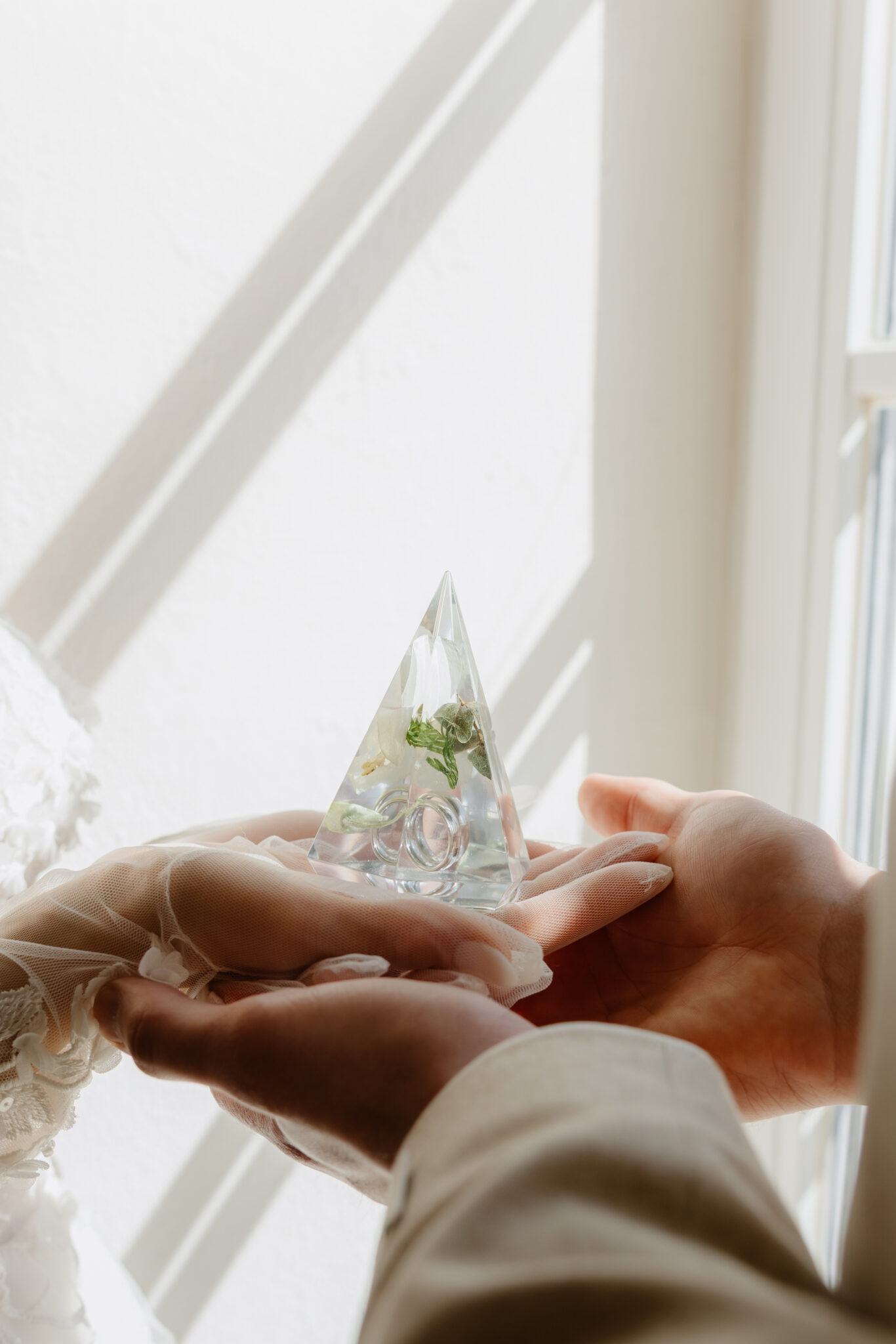 Hände halten dreieckige Acrylglas-Pyramide mit konservierten Hochzeitsblumen und Eheringen im natürlichen Licht
