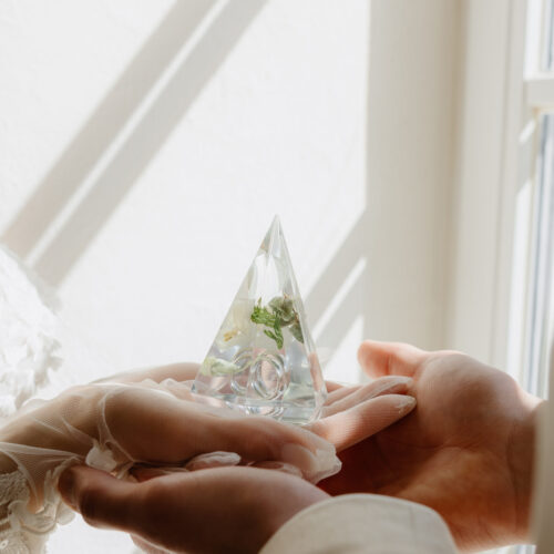 Hände halten dreieckige Acrylglas-Pyramide mit konservierten Hochzeitsblumen und Eheringen im natürlichen Licht