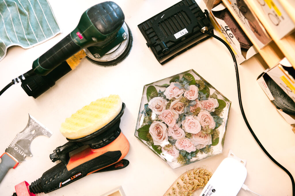 Draufsicht auf Flomory Werkstatt: Hexagonaler Resin-Block mit rosa Rosen und Eukalyptus umgeben von Schleifmaschinen, Polierscheiben und Werkzeug – Einblick in die Herstellung der Brautstrauß-Konservierung
