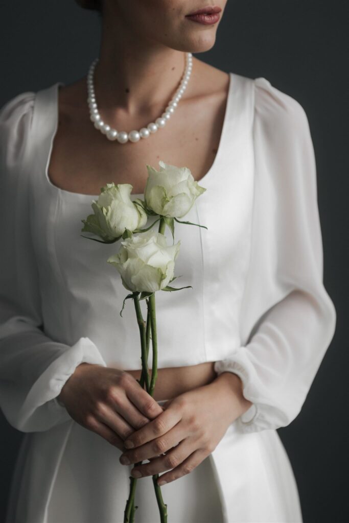 Braut im weißen Kleid mit Perlenkette hält weiße Rosen – elegantes Hochzeitsportrait – Flomory Blog