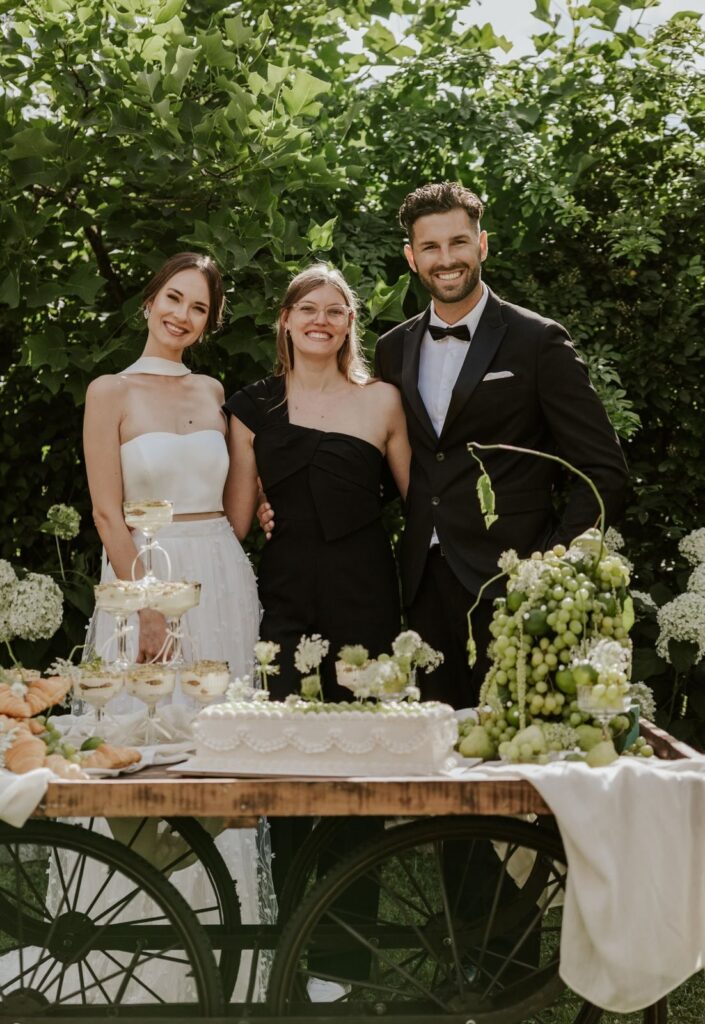 Brautpaar mit Gästin an dekoriertem Dessert-Wagen mit Hochzeitstorte und Weintrauben – Outdoor-Hochzeit im Garten – Flomory Blog