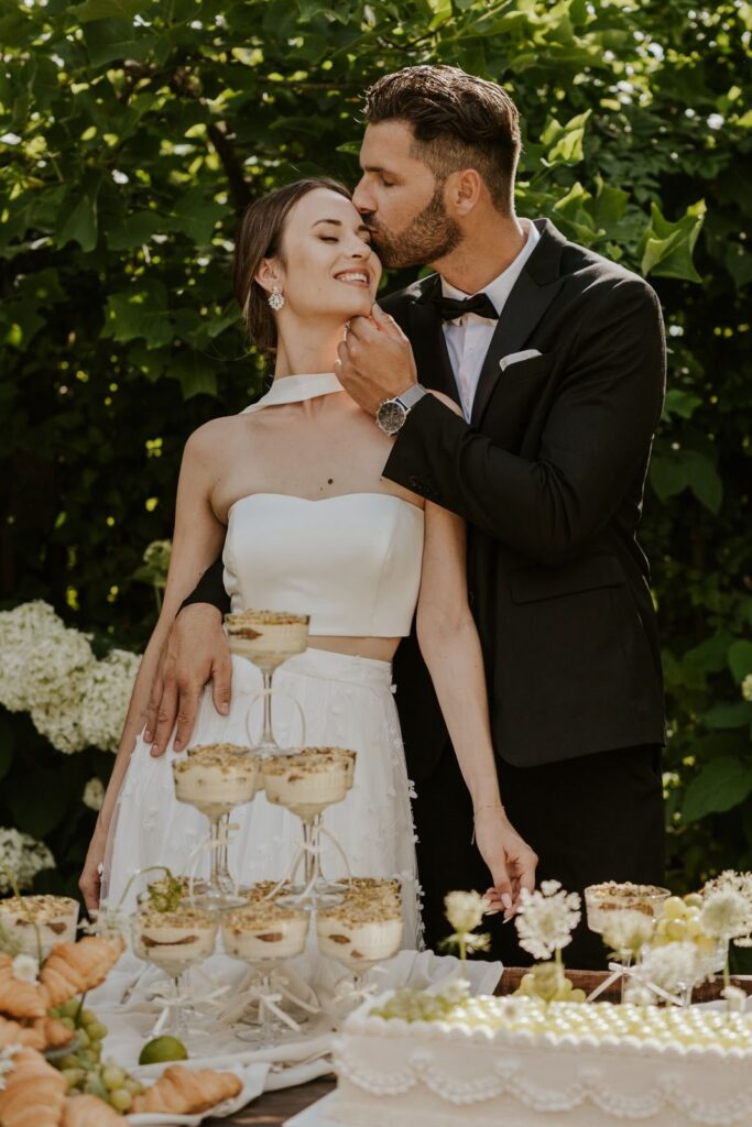 Brautpaar am Dessert-Tisch bei Outdoor-Hochzeit – Braut im weißen Zweiteiler und Bräutigam im schwarzen Smoking – Hochzeitsinspiration Flomory Blog