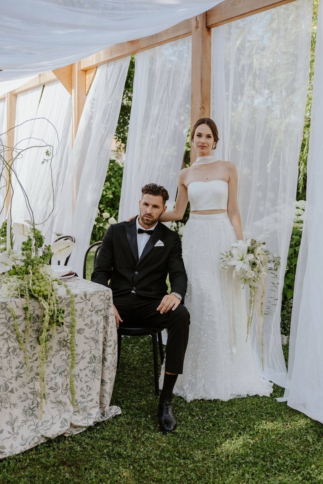 Brautpaar unter weißem Pavillon im Garten – Braut im Zweiteiler mit hängendem Brautstrauß, Bräutigam im schwarzen Smoking – Hochzeitsinspiration Flomory Blog